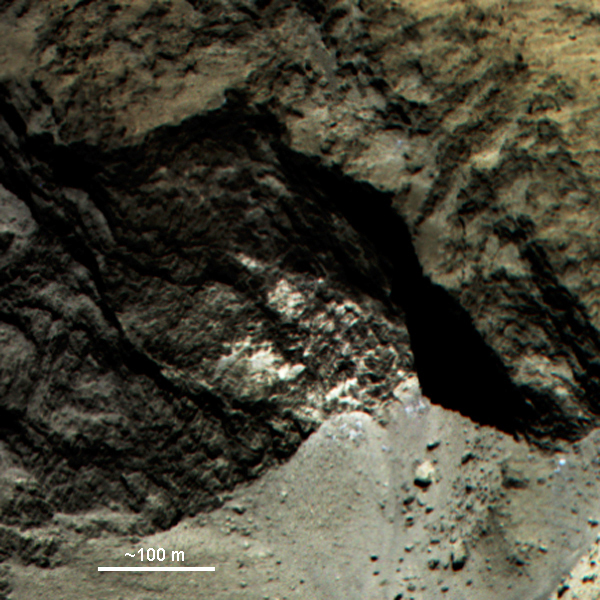 Farbkomposit eines Teils von 67P. Am Boden unterhalb der Felswand ist Material sichtbar, das heller und blauer ist als die Umgebung. Dabei könnte es sich um Blöcke (blauen) Wassereises handeln.