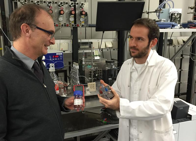 Prof. Elias Klemm (l.) und Doktorand Dennis Kopljar (mit Mikroreaktor in der Hand) vor der Anlage zur elektrochemischen Umsetzung von CO2