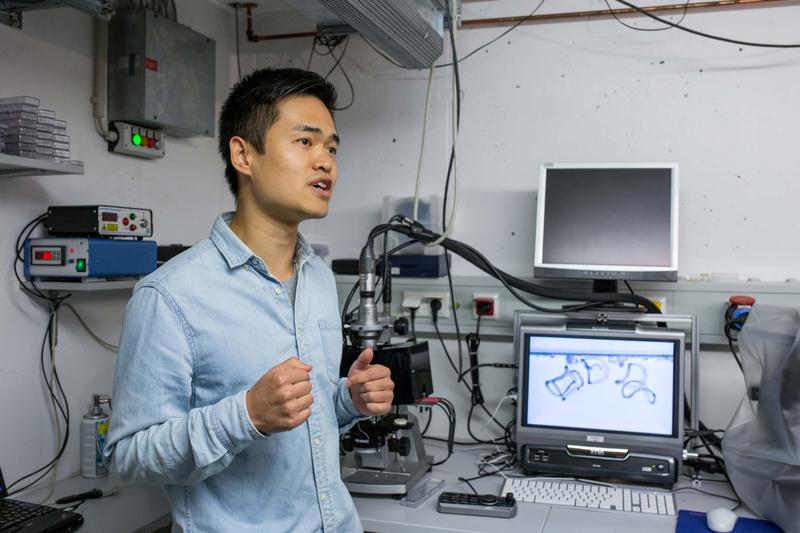 Doktorand Hang Zhang steht vor dem Mikroskop, mit dem er winzige Gel-Strukturen untersucht, die sich wie kleine Roboter selbstständig im Wasser bewegen können.