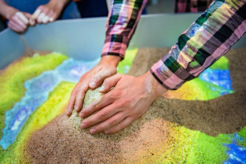 Aus Sand kann im Labor ein Höhenprofil aufgeschichtet werden. Ein Scanner erfasst es und projiziert farbcodierte Höhenlinien darauf. Mit Handbewegungen kann virtueller Regen ausgelöst werden.