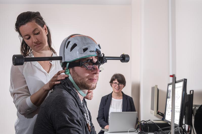 Dr. Cecilia Ramaioli (links) überprüft den Sitz des Helms mit Gewichten bei einem Probanden. Prof. Nadine Lehnen stellt im Hintergrund den Computer für das Experiment ein. 