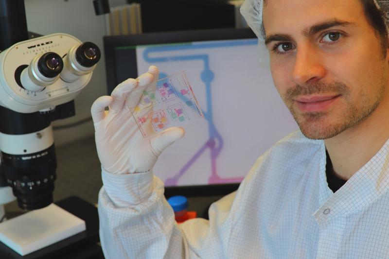 Anthony Beck, Mit-Entwickler der Technologie, hält einen chemischen Schaltkreis mit gefärbten Analysemedien. Im Hintergrund ist ein Mikroskopiebild eines der chemischen Transistoren des IC zu sehen.