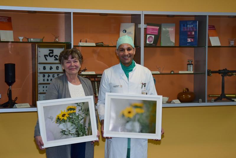 Patientin Jutta Poisel und Oberarzt Prof. Raiskup halten zwei Bilder der Fotoausstellung in den Händen. Die linke Aufnahme zeigt, wie verschwommen die Sicht bei degenerierter  Hornhautschicht ist.