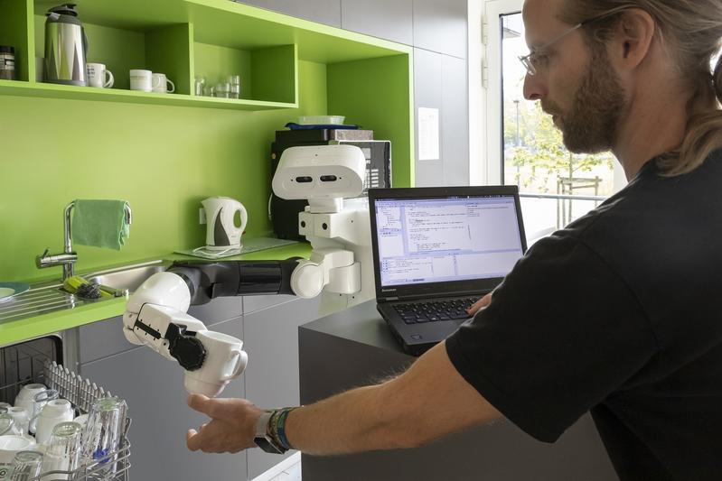 Roboter Tiago reicht in einer Küche des DFKI in Osnabrück einem Wissenschaftler eine Tasse aus der Spülmaschine.