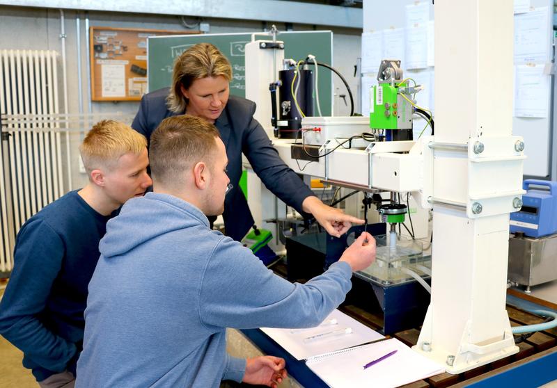 Prof. Dr.-Ing. Petra Maier erklärt Studierenden an der Werkstoffprüfmaschine das Versuchsschema.
