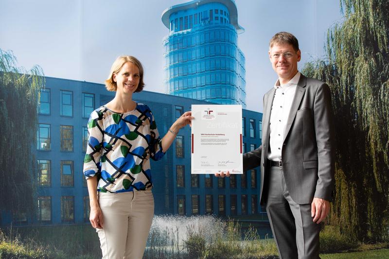 Janna von Greiffenstern, Pressereferentin und Michael Knöthig, Verwaltungsleiter an der SRH Hochschule Heidelberg, halten das Zertifikat "Familienfreundliche Hochschule" in der Hand.