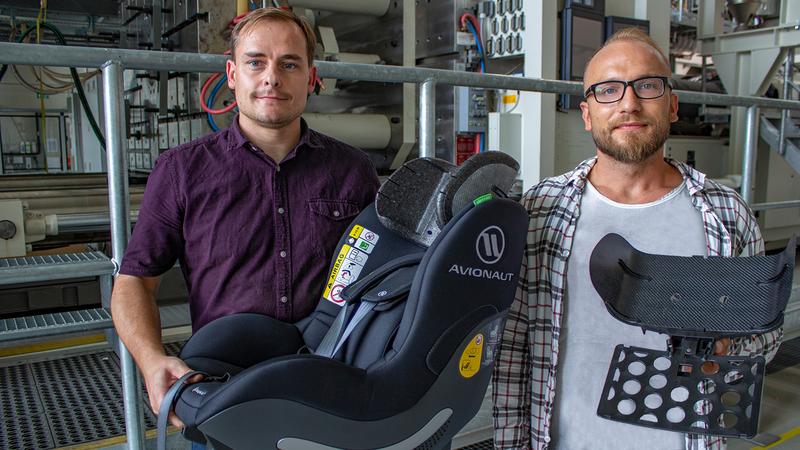 Die TU-Forscher Norbert Schramm (l.) und Tomasz Osiecki zeigen in der Halle des MERGE Technologiezentrums der TU Chemnitz den fertigen Kindersitz mit eingebauter Kopfstütze sowie die dort hergestellte Organoblech-Preform mit Trägerplatte.