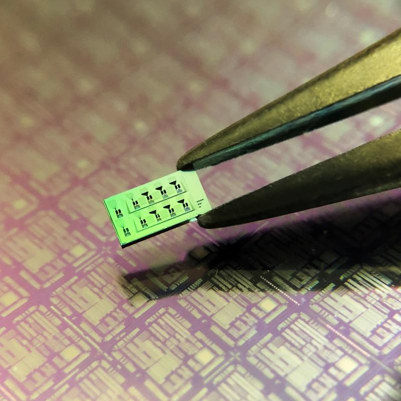Siliziumchip mit mehreren Detektoren. Die feinen schwarzen Gravuren auf der Oberfläche des Chips sind die optischen Schaltkreise, die die Detektoren miteinander verbinden (mit bloßem Auge nicht sichtbar). Im Hintergrund eine größere optis