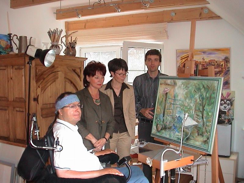 Der Mundmaler Thomas Kahlau in seinem Atelier an der neuen steuerbaren Staffelei mit der Präsidentin der Fachhochschule Lausitz, Brigitte Klotz, der Bürgermeisterin der Gemeinde Schwielowsee, Kerstin Hoppe, und dem Projektkoordinator der Hochschule, Ingo Karras (v. li.).