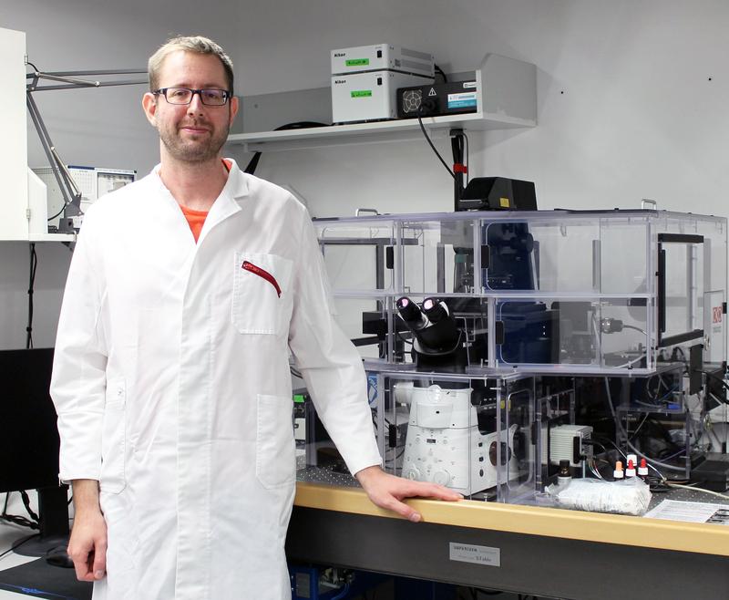 Dr. Daniel Pfeiffer in einem Labor der Bayreuther Mikrobiologie neben einem 3D-SIM-Mikroskop, das fluoreszenzmikroskopische Aufnahmen mit hoher Auflösung ermöglicht.