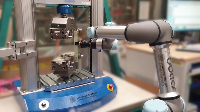 Der kollaborierende Roboter legt eine Materialprobe in die Zugprüfmaschine.  Im Projekt werden weitere Messeinrichtungen wie z.B. Kameras für die automatisierte Versuchsdurchführung integriert. 
