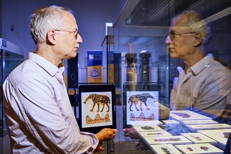 Prof. Dr. Ludwig D. Morenz sich in einer Vitrine des Ägyptischen Museums der Universität Bonn spiegelnd. Rechts von ihm ist die Hieroglyphe für die Nilinsel Elephantine zu sehen: oben ein Elefant und unten symbolisch ein Gebirge. 