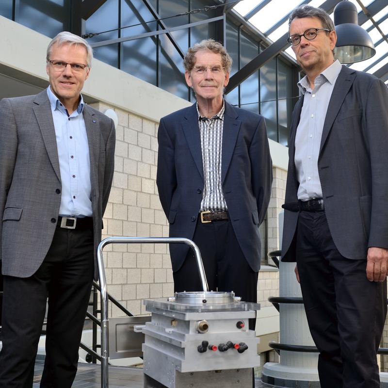 The inventors of the process, Wilhelm Meiners, Kurt Wissenbach and Anders Gasser, stand in front of a small transport trolley with the first LPBF machine.