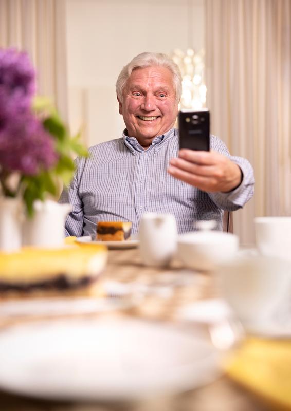 Ein Mann sitzt an einem Kaffeetisch und macht mit seinem Handy ein Selfie.