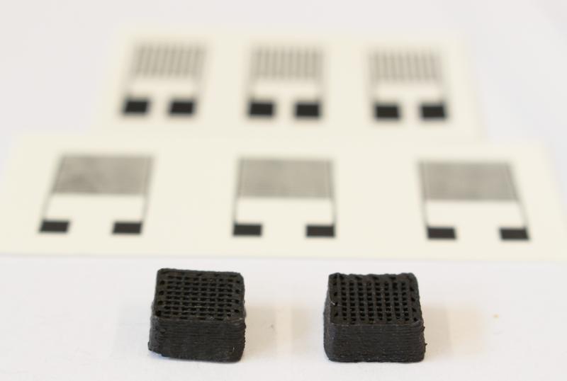 Neuartiger Werkstoff für gedruckte Schaltungen: Zwei Testquader von einem Zentimeter Breite aus dem 3D-Drucker. Im Hintergrund sieht man die gedruckten elektronischen Sensoren.