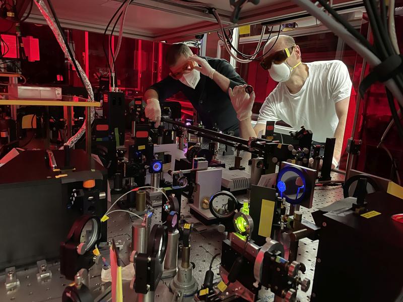 Marius Morgenroth (links) und Dr. Mirko Scholz (rechts) an der Laser-Messapparatur für die zeitaufgelöste CD-Spektroskopie, in laserschutzgerechter und Corona-konformer Ausrüstung.