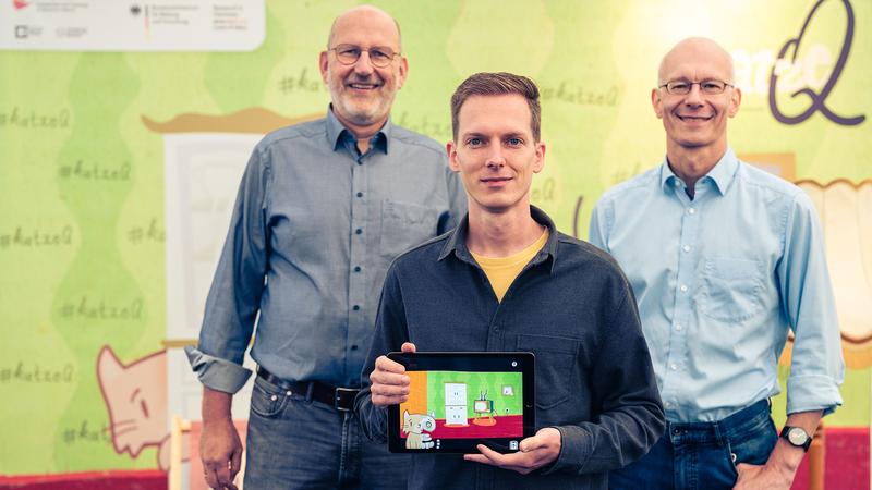 Die beiden Sprecher des Exzellenzclusters ct.qmat, Prof. Ralph Claessen (l.) und Prof. Matthias Vojta (r.), präsentieren gemeinsam mit dem App-Designer Philipp Stollenmayer (Mitte) ihr gemeinsames Projekt, die Spiele-App „Katze Q“.