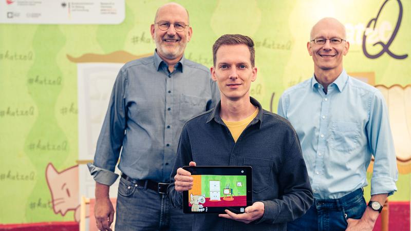 The two spokespersons of the Cluster of Excellence ct.qmat, Prof. Ralph Claessen (l.) and Prof. Matthias Vojta (r.), together with app designer Philipp Stollenmayer (center), present their joint project, the game app "Kitty Q".