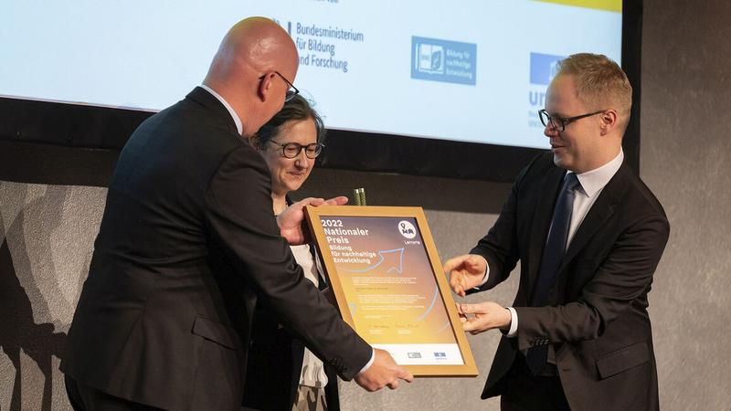 Johannes Orphal (links), Beauftragter des Präsidiums für Klimaschutz und Nachhaltigkeit, und Senja Post, wissenschaftliche Leiterin des ZAK nehmen den „Nationalen Preis – Bildung für nachhaltige Entwicklung“ von Staatssekretär Jens Brandenburg entgegen