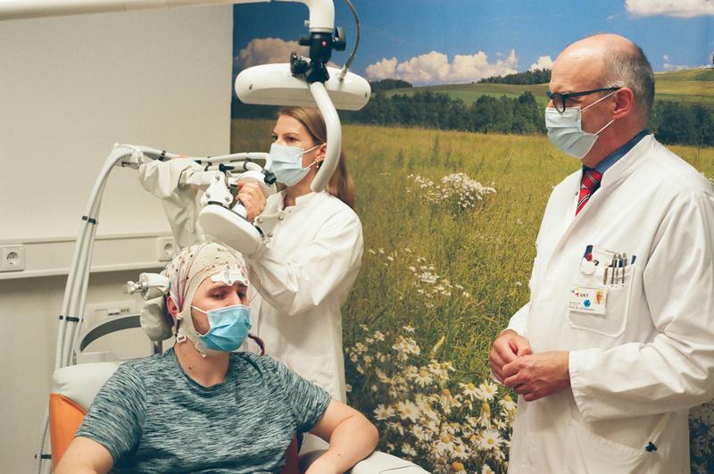 Personalisierte Hirnstimulation eines gesunden Probanden. Eine Magnetspule stimuliert sein Gehirn von außen während gleichzeitig der momentane Erregungszustand mit einem EEG gemessen wird. Rechts im Bild: Prof. Dr. Ziemann.