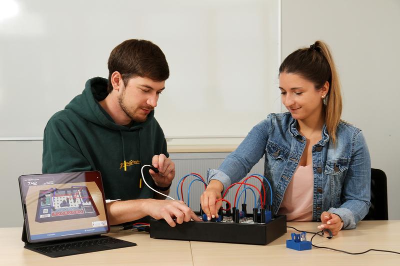 Frederik Lauer und Lena Geuer am SmaEPho. Die App auf dem Tablet spiegelt die Hardware via digitalem Zwilling. Der Aufbau (v.r.n.l): Photometrische Messeinheit, Steckbrett mit Verbindungskabeln, elektrischem Widerstand und Potentiometer, Mikrocontroller. 