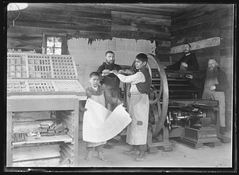 Drei Kapuzinermissionare in einer Druckerei in Valdivia, die gemeinsam mit ihren indigenen Schülern eine Druckpresse bedienen. Im Vordergrund ist ein Setzkasten mit Drucklettern zu erkennen. Die Aufnahme entstand ca. 1927.
