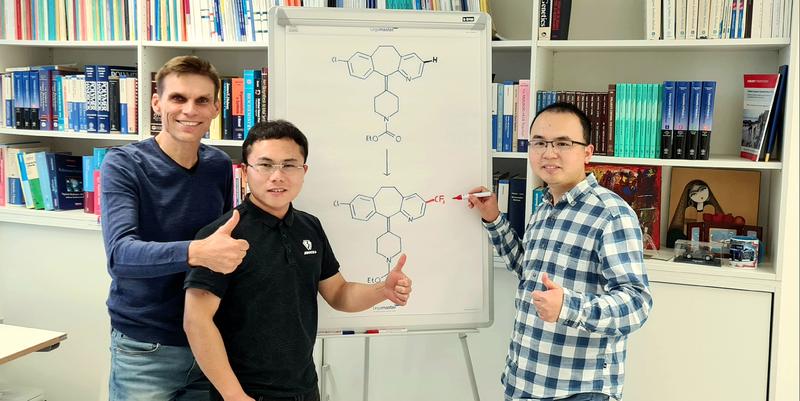 Prof. Dr. Armido Studer mit Dr. Hui Cao und Dr. Qiang Cheng (v. l.). Auf der Tafel ist die chemische Transformation von Loratadin dargestellt, einem wichtigen Histamin-H1-Rezeptor-Antagonisten, welcher durch eine Trifluormethylgruppe modifiziert wurde.