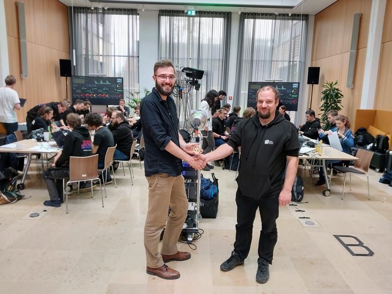 Prof. Dr. Christian Dörr gratuliert Student Michael Rüßmann zur Top Platzierung. Im Hintergrund das große Attack-Defense Team Finale.