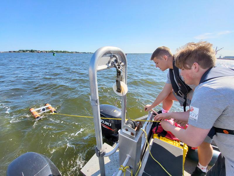 Die IESO-Olympioniken Leon Arlit und Benjamin Poost an Bord des IOW-Forschungsbootes KLAASHAHN lassen ein Messgerät mit verschiedenen Spezialsonden auf der Warnow zu Wasser, um Daten für ihr Projekt zu erheben.