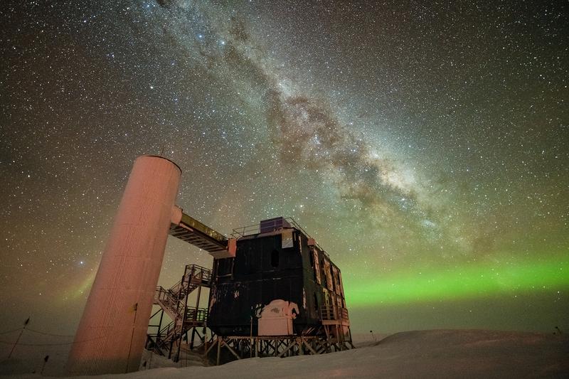 Ein Blick auf das IceCube-Labor bei einem sternenklaren Nachthimmel, der die Milchstraße und grüne Polarlichter zeigt.