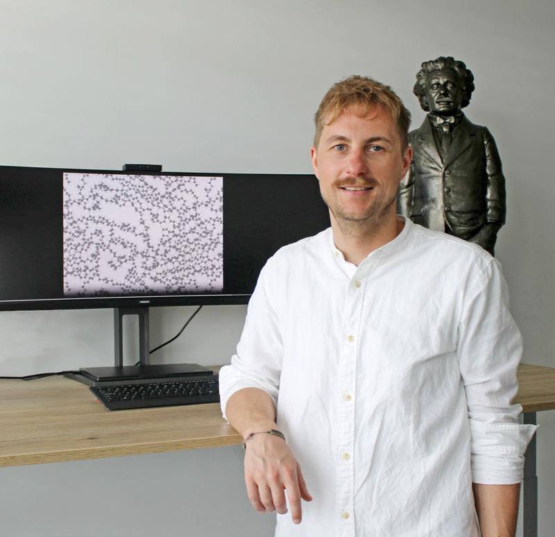 Prof. Dr. Christopher Kuenneth, Universität Bayreuth. Der Bildschirm zeigt die atomistische Struktur eines Polymers mit Kohlenstoff-Atomen (graue Kugeln) und Wasserstoff-Atomen (weiße Kugeln). Im Hintergrund eine Albert Einstein-Statue von Ottmar Hörl.
