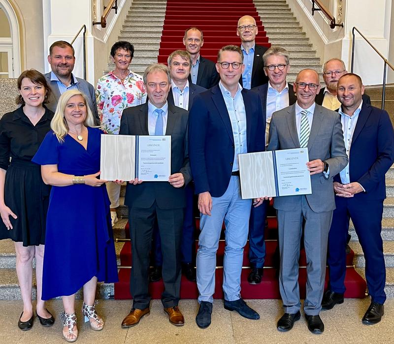 Foto: Die beiden Hochschulpräsidenten Prof. Dr. Stefan Gast und Prof. Dr. Niels Oberbeck bei der Urkundenübergabe durch Wissenschaftsminister Markus Blume – unterstützt von Repräsentant:innen der Region.