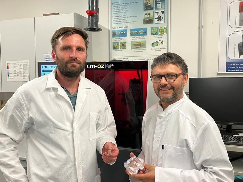 Doktorand Christian Polley (links) und Professor Hermann Seitz (rechts) (hier vor einem 3D-Drucker) untersuchen im Sonderforschungsbereich 1270 „Elektrisch Aktive ImplaNtatE – ELAINE“, wie damit große Knochendefekte behandelt werden können 