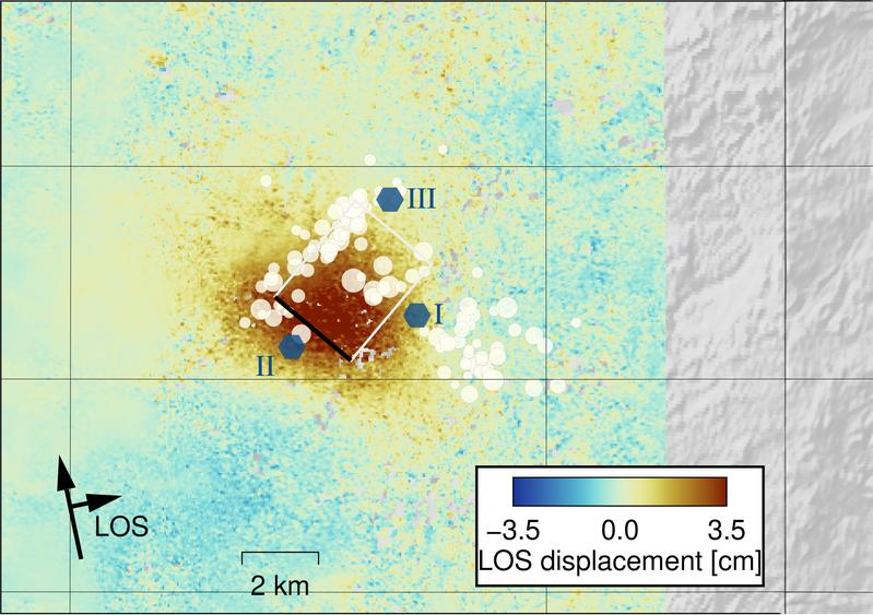 Karte der Region um Peace River im Norden der kanadischen Provinz Alberta, Kanada. Der farbige Hintergrund stellt die durch Satelliten gemessene Oberflächenverformung dar. Weiße Kreise: Erdbeben; blaue Sechsecke: Injektionsstellen der Industrieabwässer.