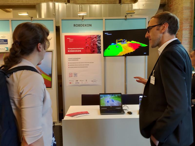 Janko Petereit zeigt am ROBDEKON-Stand beim BMBF-Innovationsforum die Interaktion mit dem autonomen Bagger ALICE. Über die Monitore ließ sich der Bagger beobachten und die Fässer auswählen.