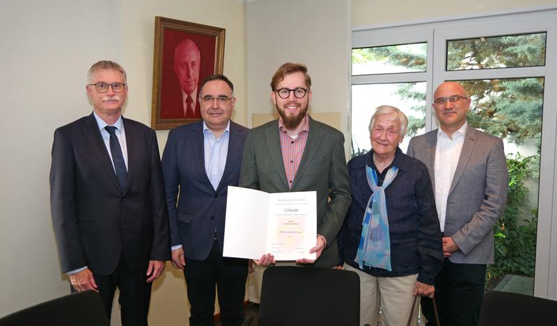 Preisträger Jakob Halfmann mit dem Vorstand der Ruchti-Stiftung und deren Beiratsmitglied (v.l.) Unikanzler Dr. Uwe Klug, Professor Ignacio Czeguhn, Gertrud Schrödl und André Illmer. Das Foto an der Wand zeigt den Stiftungsgründer Wilhelm H. Ruchti.