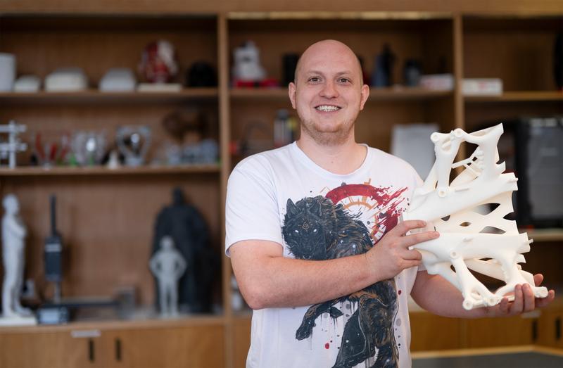 Felix Felgenträger zeigt einen 3D-gedruckten Vakuumgreifer. Im Regal im Hintergrund stehen weitere Exponate aus dem 3D-Druck-Spektrum der Hochschule  Coburg wie die bekannte 3D-gedruckte Coburger Prinz-Albert-Statue.