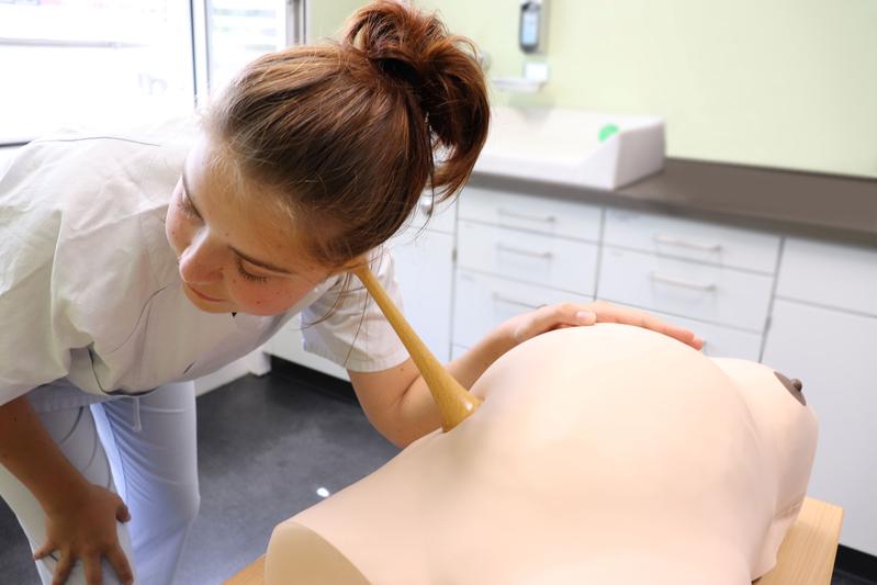 Eine Studentin in medizinischer Kleidung hört mit einem hölzernen Stethoskop die Herztöne eines Schwangerschaftssimulators ab.