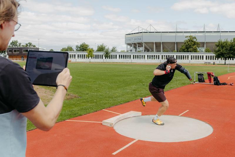 Paraathlet Alex Bartz stößt mit der neu entwickelten Kugelstoßkugel. Projektmitarbeiter Dan Bürger beobachtet den Stoß über die App auf dem Tablet.