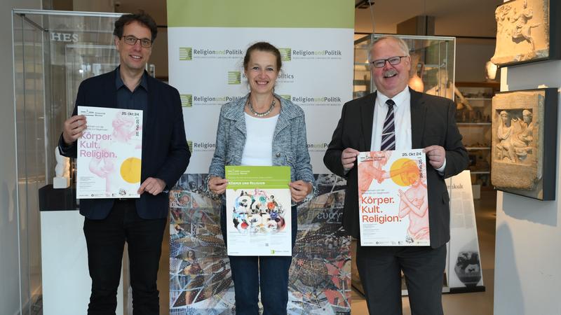 Exhibition organizers Achim Lichtenberger (Archaeology), Angelika Lohwasser (Egyptology), and Holger Strutwolf (Theology) with the exhibition posters and the poster for the annual theme "Religion and Body"