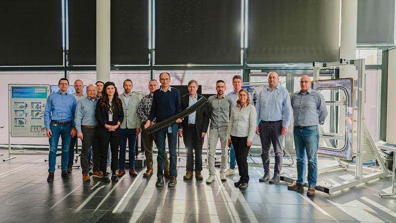 Das TAVieDA-Projektteam: Christian Wolf, Projektleiter bei Airbus, präsentiert einen Randträger aus Thermoplast-Verbundmaterial. 4. von rechts: Dirk Herborg (DLR Projektträger); im grauen Sakko: Dr. Rayk Fritzsche. 5. von links: Maxi Grobis. 
