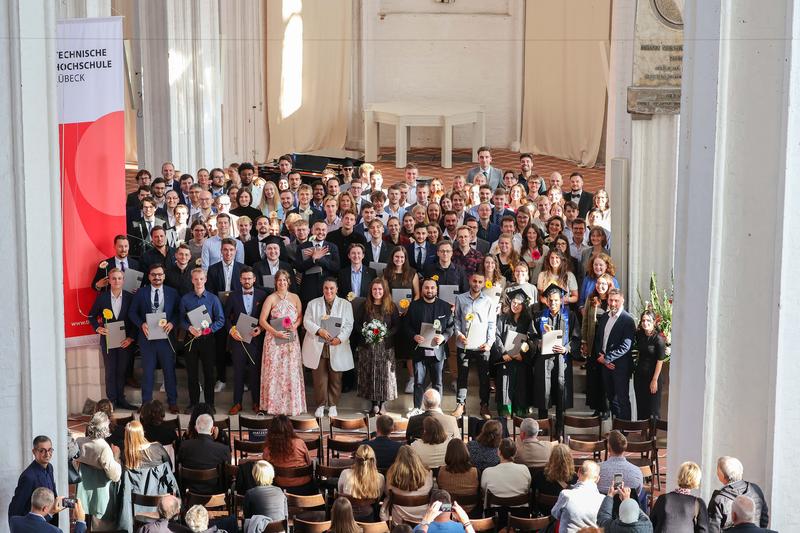 Die Absolvent*innen der TH Lübeck auf einem Foto. Viele halten Urkunden und Blumen, Gäste sitzen im Vordergrund. 
